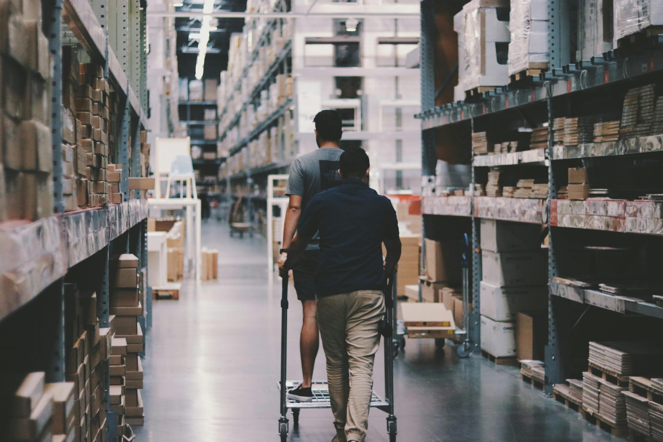 pexels photo 1797428 1797428 Two men maneuver a trolley in a large warehouse filled with boxes and shelves.